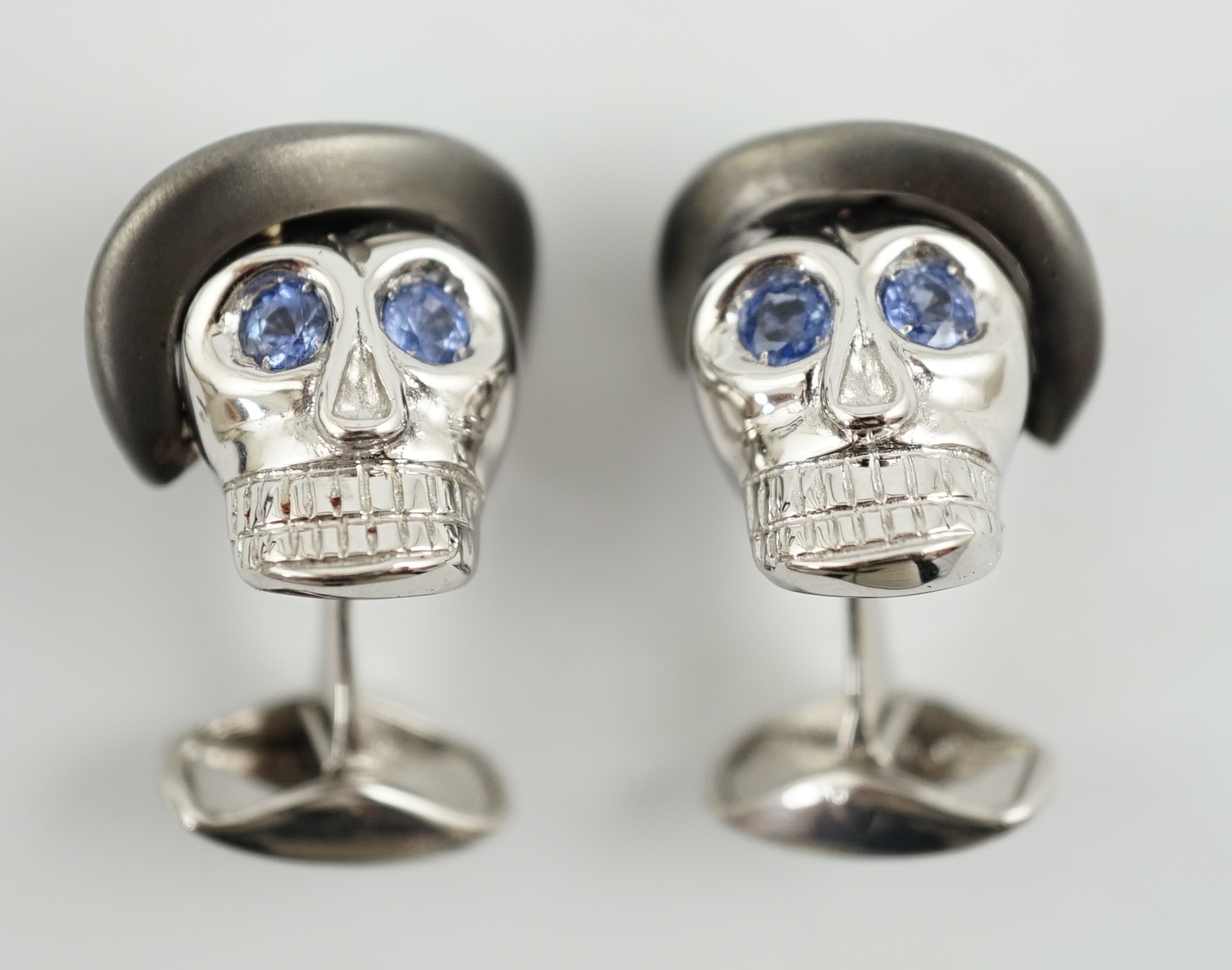 A modern pair of Longmire 18ct white gold, blackened rhodium and sapphire set novelty cufflinks, each modelled as a skull wearing a bowler hat